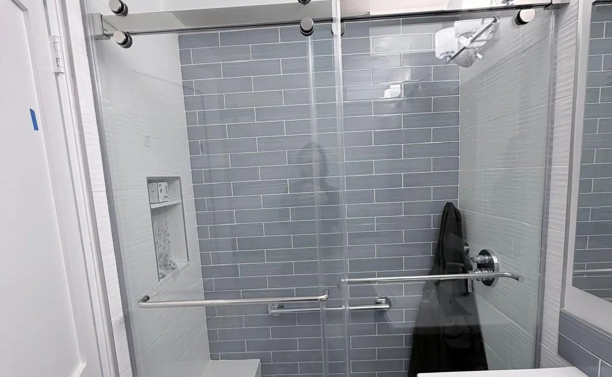 Sliding shower enclosure with an overhead rail, round rollers and long horizontal handles in front of gray wall tile.