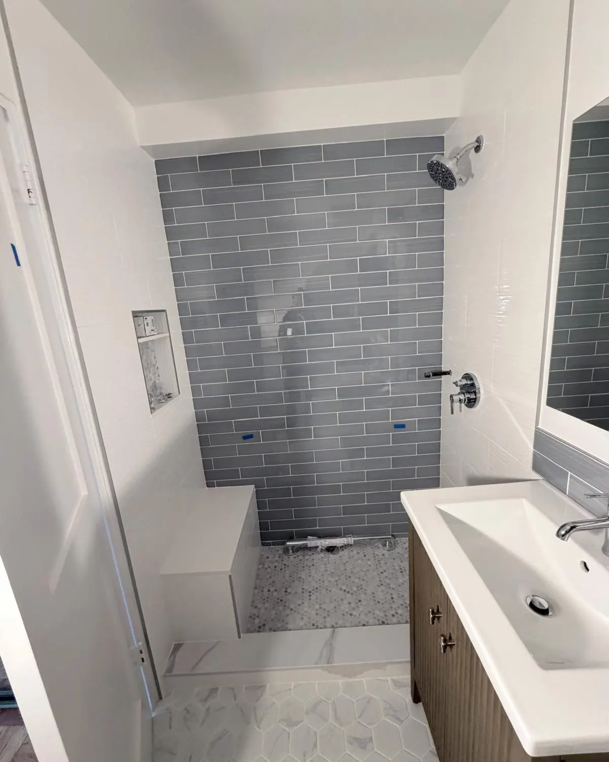Shower opening before glass at 84 Clinton, with gray back-wall tile, a pale bench and curb, fixtures and a nearby vanity.