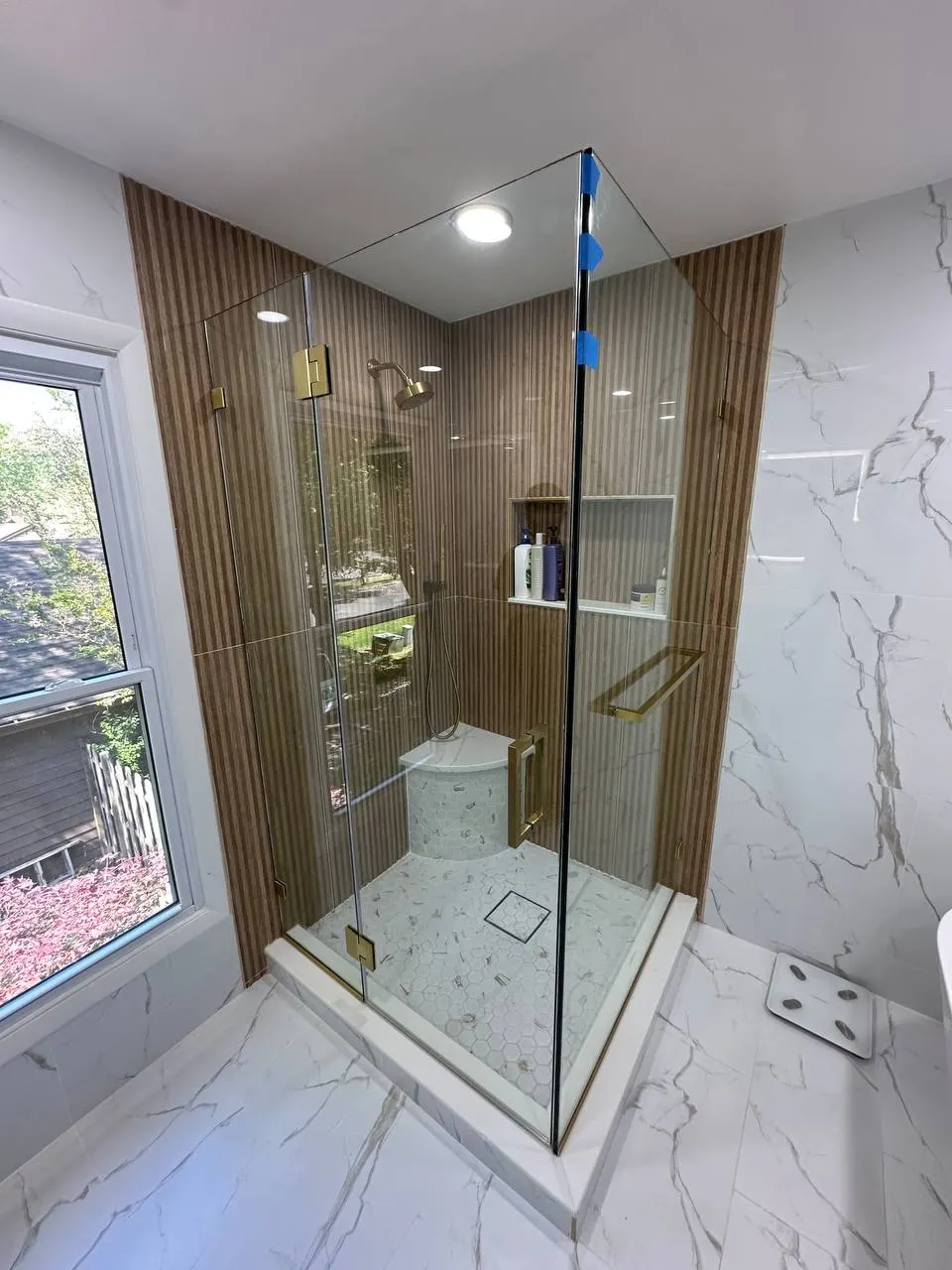 Clear corner shower enclosure with brass-colored hinges beside vertically striped brown tile and a pale curb.