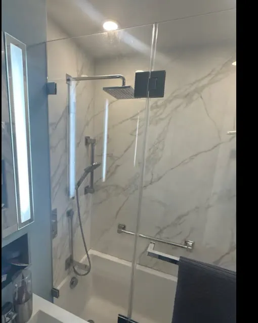 Frameless tub enclosure with chrome hardware and rain shower head against white marble tile, remediated NYC project