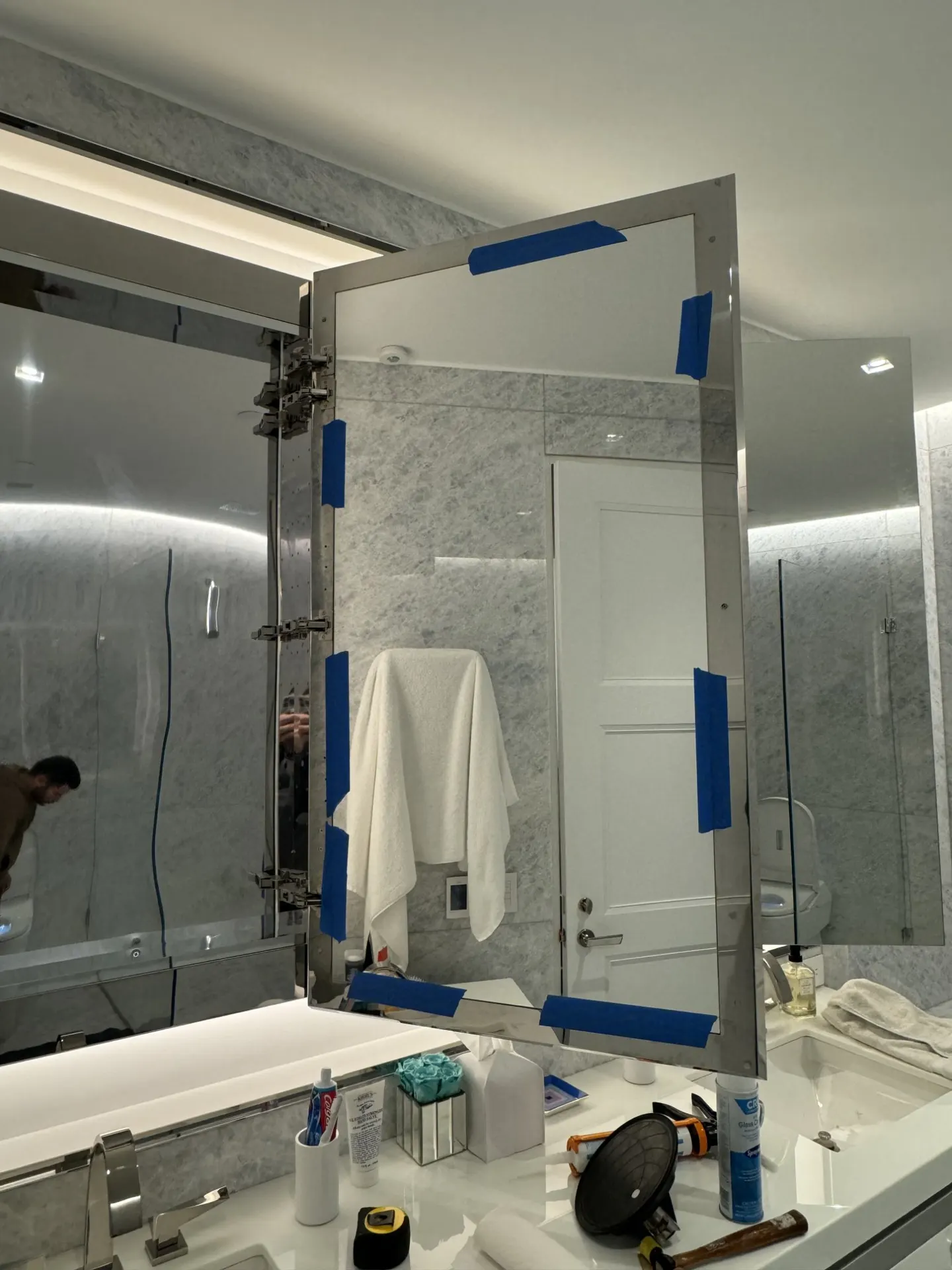 Open mirrored cabinet door with blue tape at its edges, visible hinges and reflected bathroom lights beside shower glass.